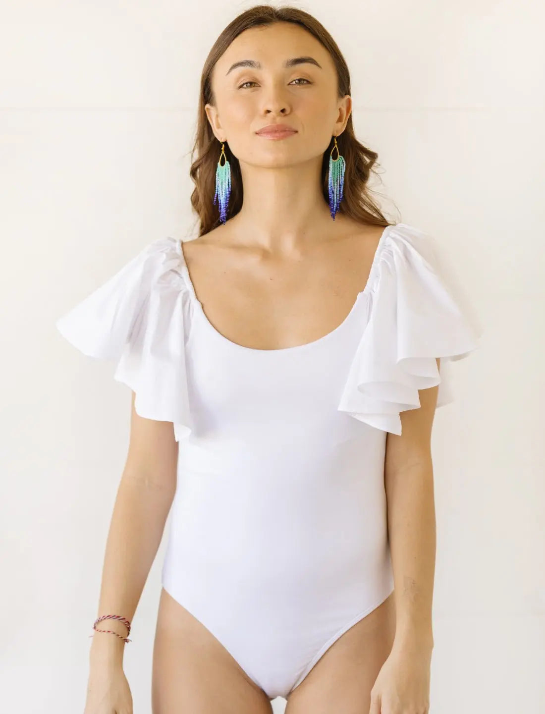 Model wears a white bodysuit with flutter sleeves and a scoop neckline. The bodysuit has ruffled details on the shoulders and a form-fitting silhouette. 