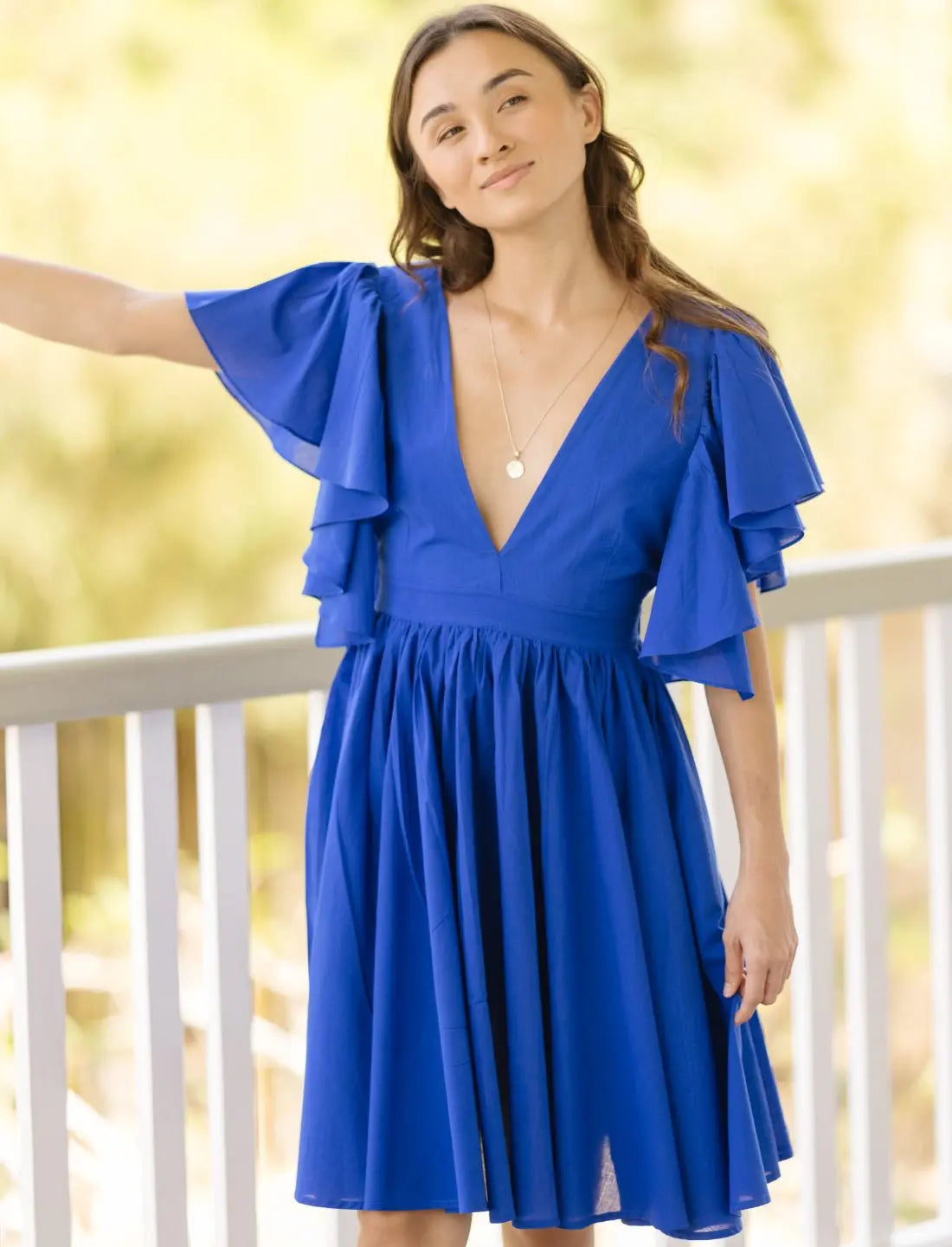 Playful Ina Short Dress in royal blue featuring cascading ruffle sleeves, plunging v-neckline, and flowy gathered skirt. Summer cocktail dress, feminine silhouette, vacation outfit, wedding guest attire, ethical fashion, sustainable clothing, bohemian inspired design, romantic daywear, garden party dress, handcrafted apparel, resort wear, photographed in soft natural lighting on outdoor balcony.