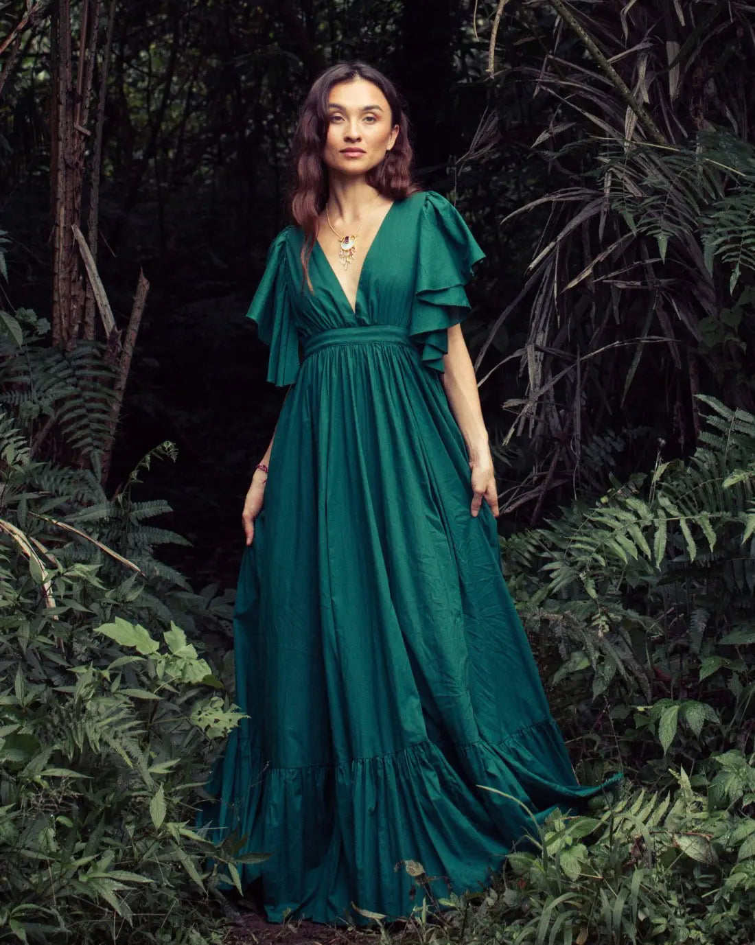 Versatile Spirit Maxi Dress in emerald green with flutter sleeves, styled on a model in a lush tropical forest setting. Bohemian fashion, flowy silhouette, ethical clothing, sustainable fashion, forest photoshoot, nature-inspired design, v-neck dress, statement jewelry, eco-friendly apparel, artisan crafted, festival outfit.