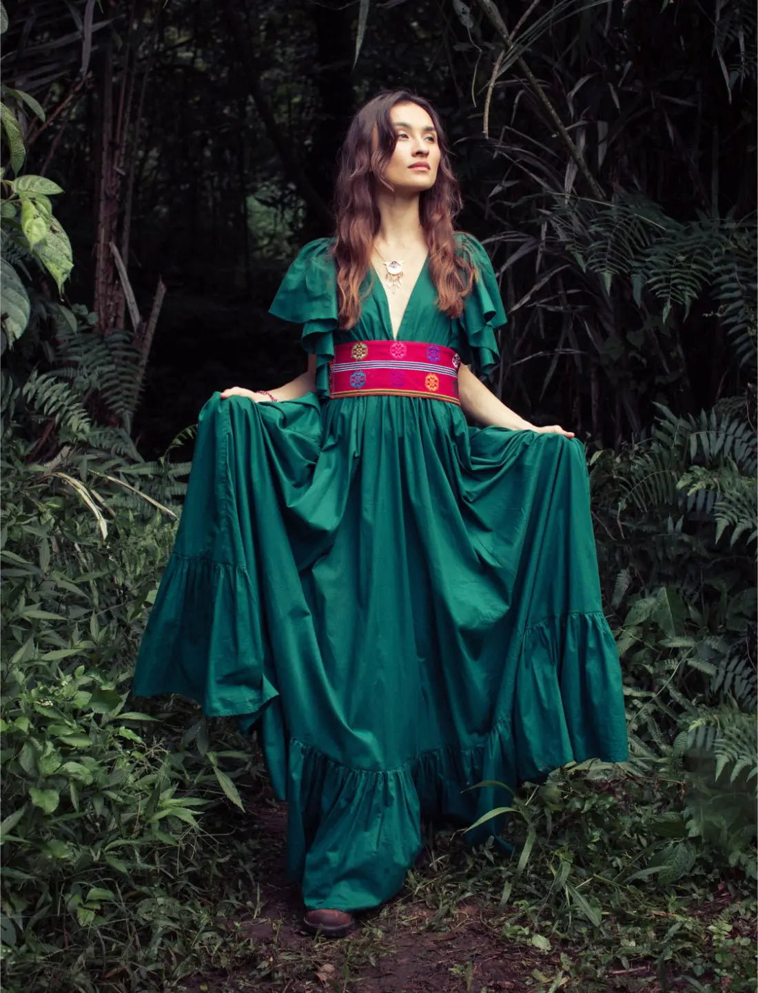 Versatile Spirit Maxi Dress in emerald green with flutter sleeves and colorful embroidered belt, styled on a model in a lush tropical forest setting. Bohemian fashion, flowy silhouette, ethical clothing, sustainable fashion, forest photoshoot, nature-inspired design, v-neck dress, statement jewelry, eco-friendly apparel, artisan crafted, festival outfit.