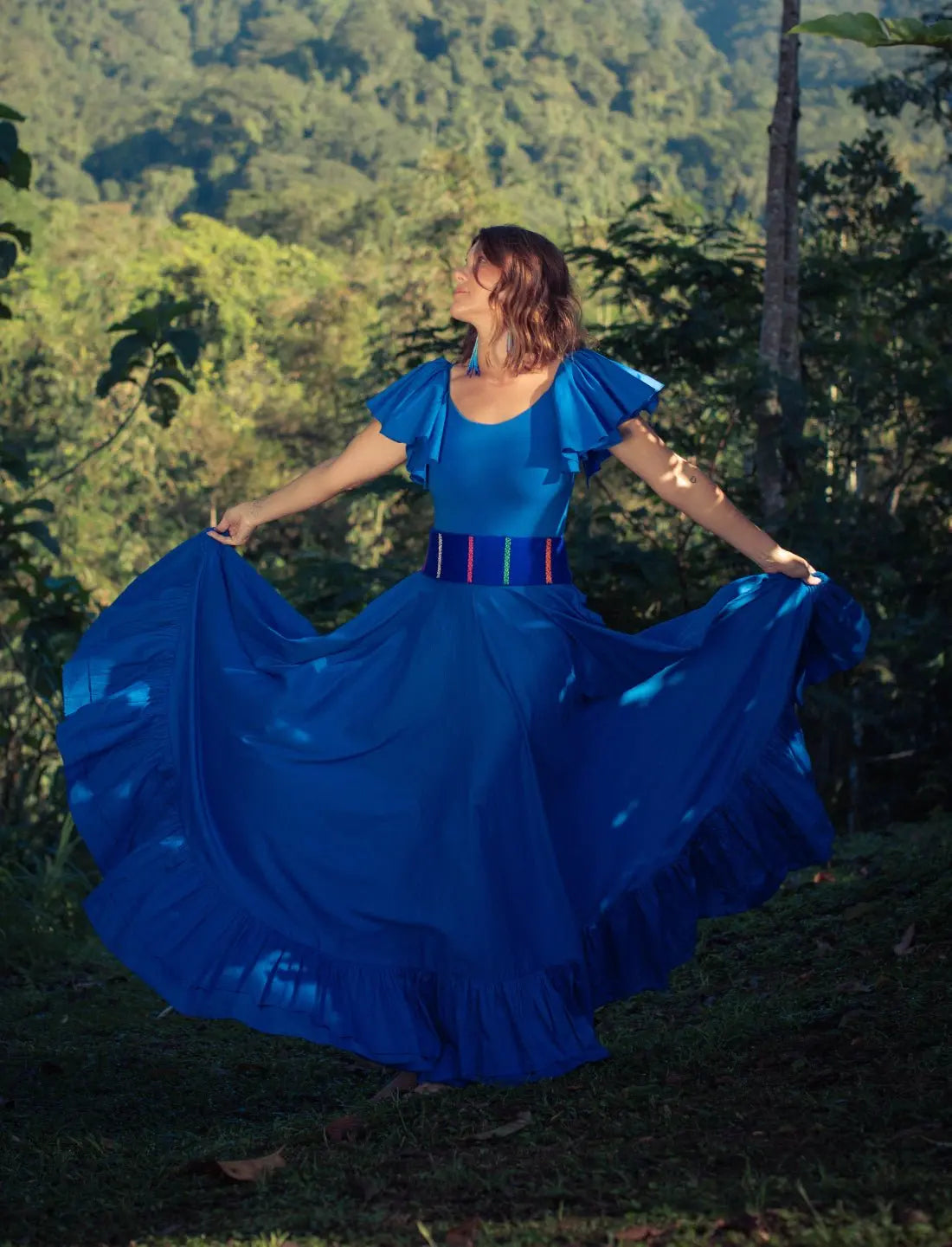 Sacred Hoop of Life Maxi Skirt in royal blue shown in full twirl with matching flutter sleeve top and Colors of the Cosmos Belt. Bohemian maxi dress in motion, ethical fashion, sustainable clothing, mountain backdrop, ruffle hem design, dancing silhouette, handcrafted apparel, vibrant blue dress, artisan accessories, festival fashion, outdoor photoshoot, nature-inspired style photographed against lush jungle mountains.