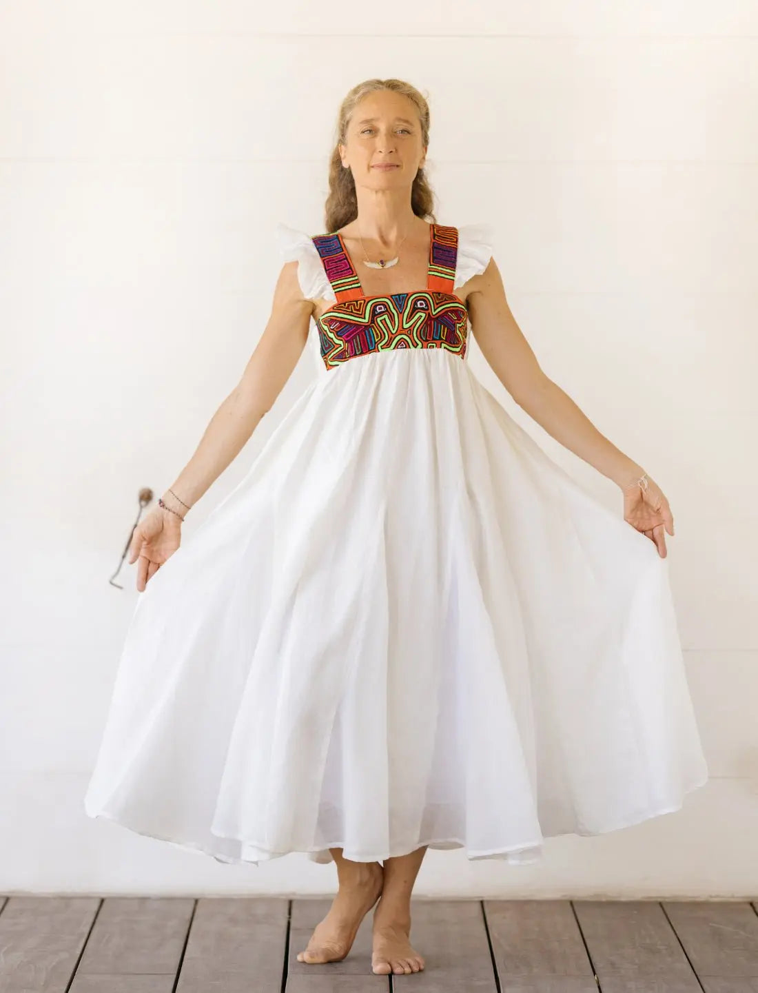Model wearing the Yemaya Embroidery Adult Gown in white linen with colorful geometric mola embroidery at the bodice. The mola pattern includes two colorful pelicans on the front. The flowing gown features flutter sleeves and a loose, comfortable silhouette. Model is showing the dress's movement by holding out the skirt against a neutral background.