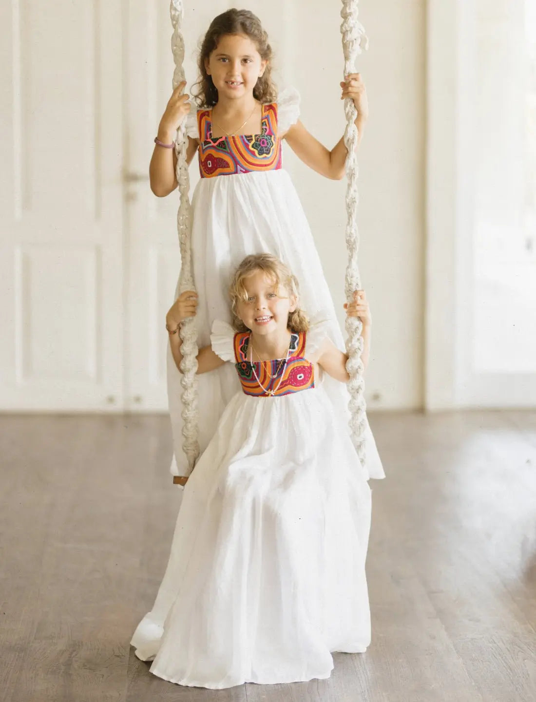 Two young girls wearing matching Yemaya Child Gowns in white linen with colorful mola embroidery on the bodice. The girls are sitting and standing on a rope swing indoors, both smiling at the camera. The flowing white dresses feature vibrant orange and red geometric patterns at the neckline, showcasing the traditional Kuna handicraft. The gowns demonstrate their generous fit and playful, twirl-worthy silhouette perfect for children's movement and play.