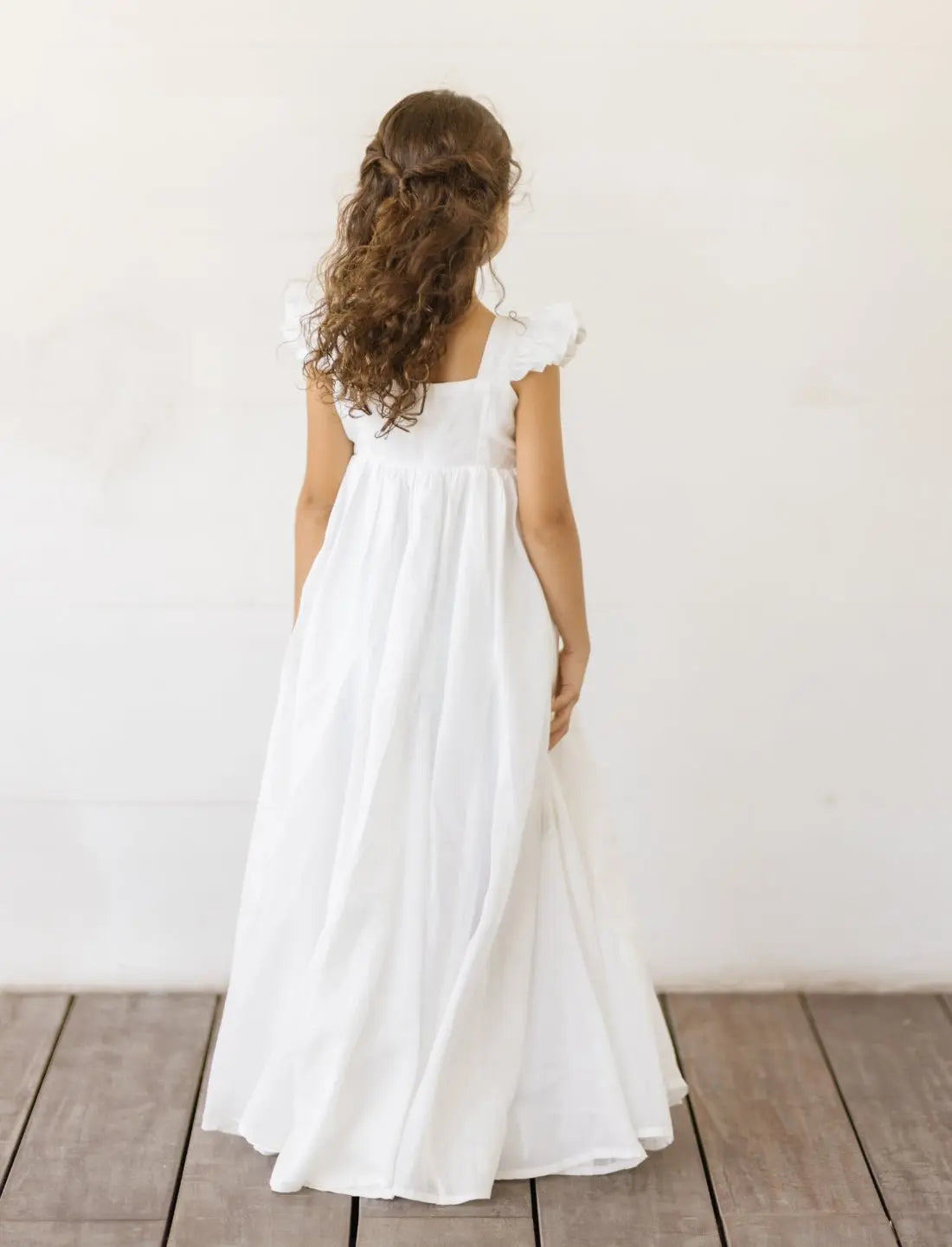 A young girl turns away from the camera, showcasing the solid white linen backside of the Yemaya Embroidery Child Gown. The dress drapes beautifully, with elegant flutter cap sleeves. Versatile and comfortable, this gown is a perfect addition to your child's wardrobe.