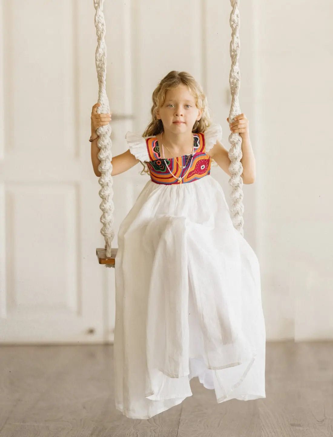 Young girl with blonde curly hair sitting on a rope swing, wearing the Yemaya Child Gown. The white linen dress features colorful mola embroidery on the bodice with vibrant orange, red, blue, and black geometric patterns created by Kuna artisans. The gown shows off its delicate flutter sleeves and flowing design as she holds the rope swing against a neutral background. The airy fabric drapes beautifully, demonstrating the dress's movement and playful nature perfect for children's activities.