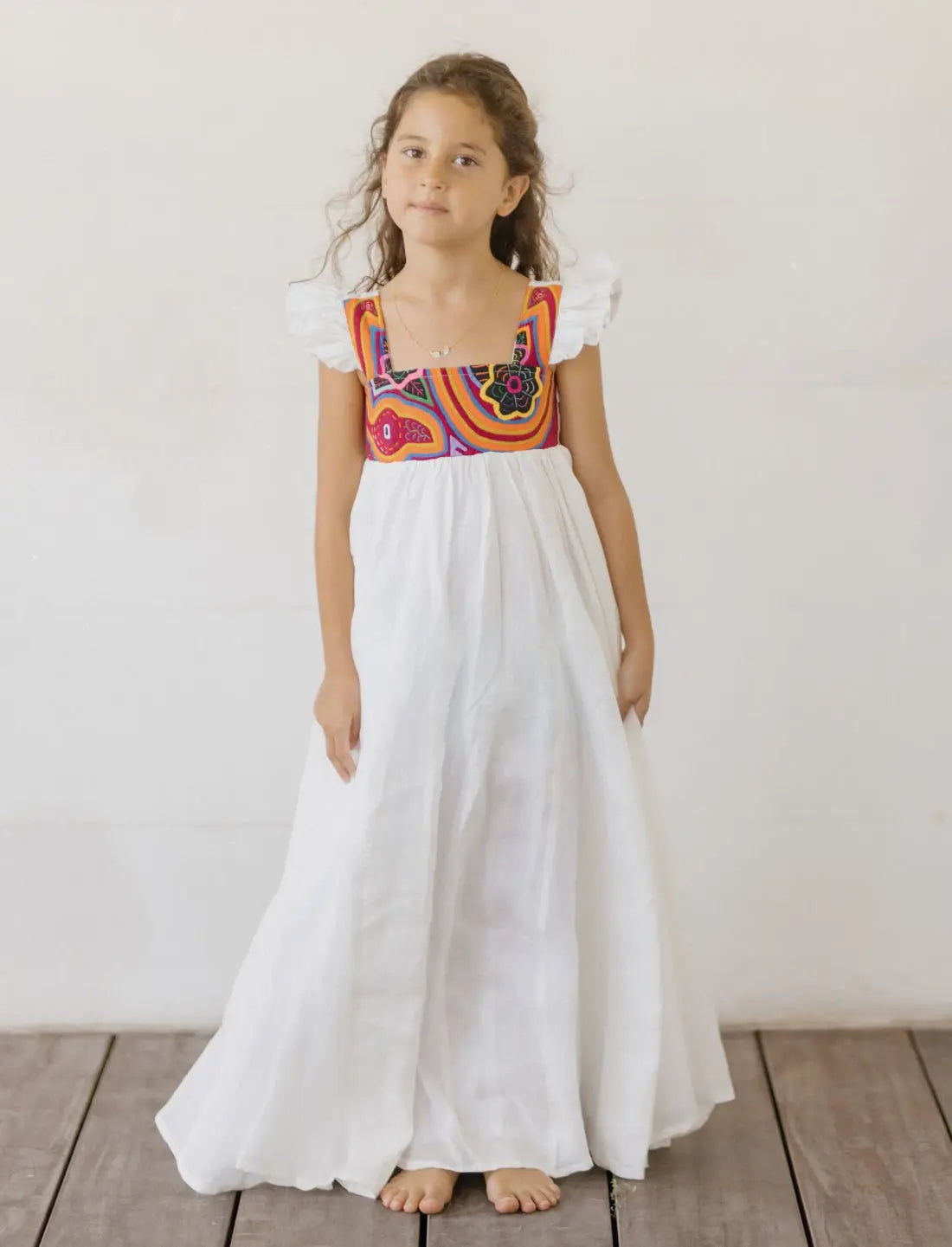 A young girl wears a full-length Yemaya Child Gown against a neutral background with wooden flooring. The white linen dress features colorful mola embroidery at the bodice with vibrant red, orange, blue, and pink geometric patterns made by Kuna artisans. The gown showcases its flutter cap sleeves with ruffle detail and flowing floor-length silhouette. The child stands barefoot, demonstrating the generous fit and elegant drape of the premium linen fabric.